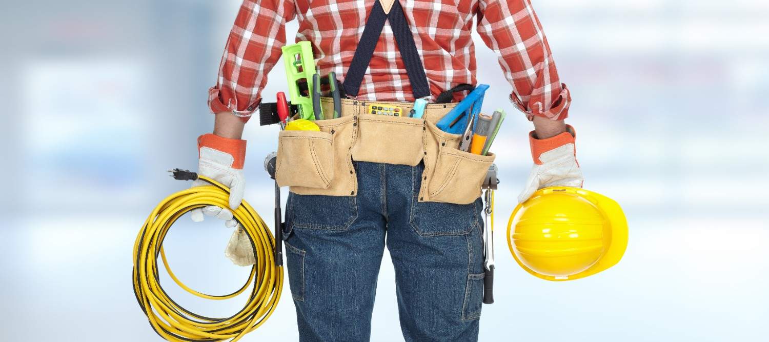 Tradesperson wearing a loaded tool belt, holding a yellow extension cord and hard hat, ready for jobsite work.