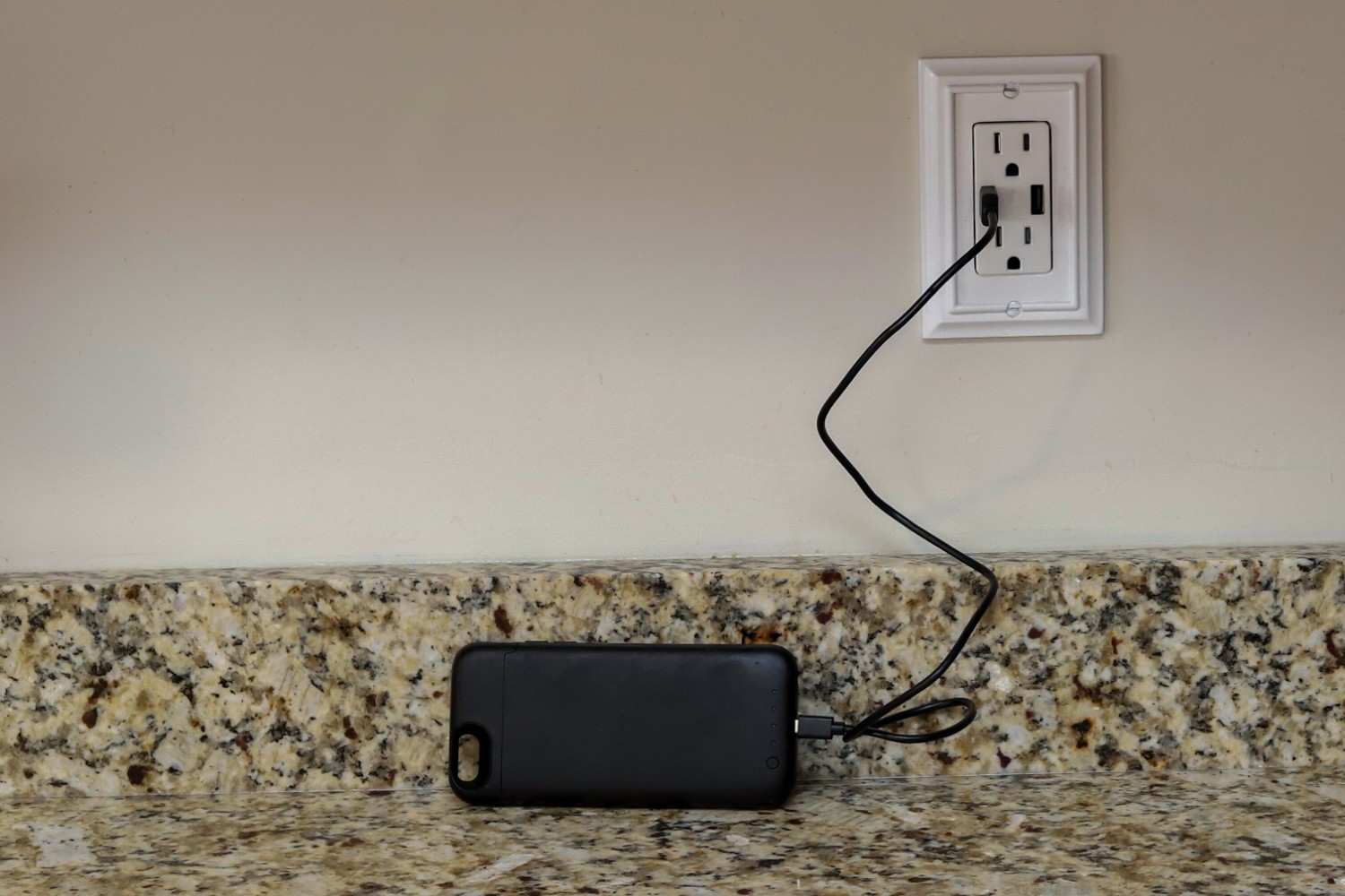 Smartphone charging directly from a wall outlet with built-in USB ports on a kitchen counter, demonstrating adapter-free power access.