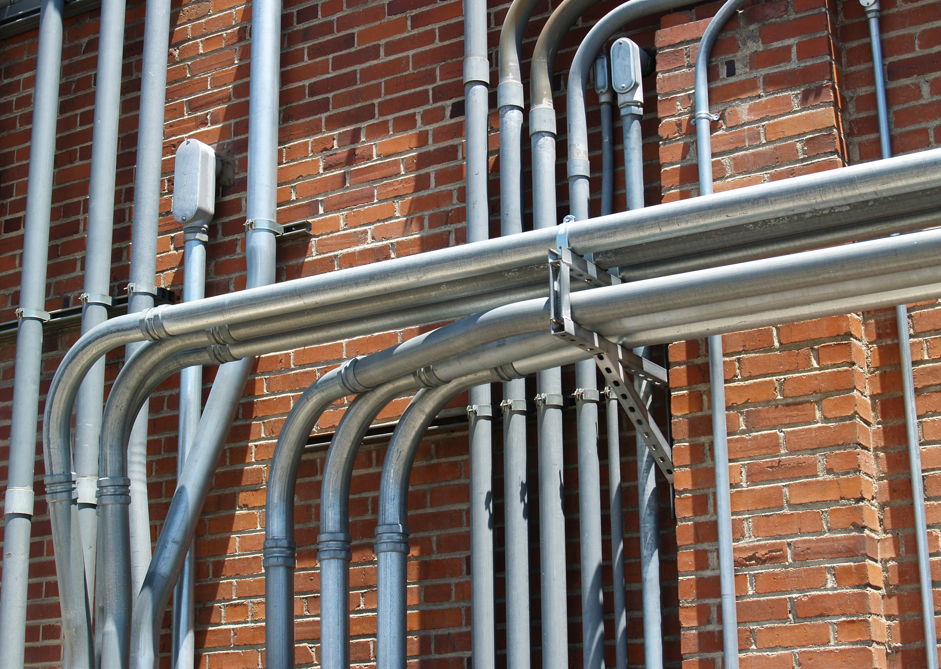 Mounted metal conduit network with connected fittings and junction boxes installed on a brick exterior of a commercial building.